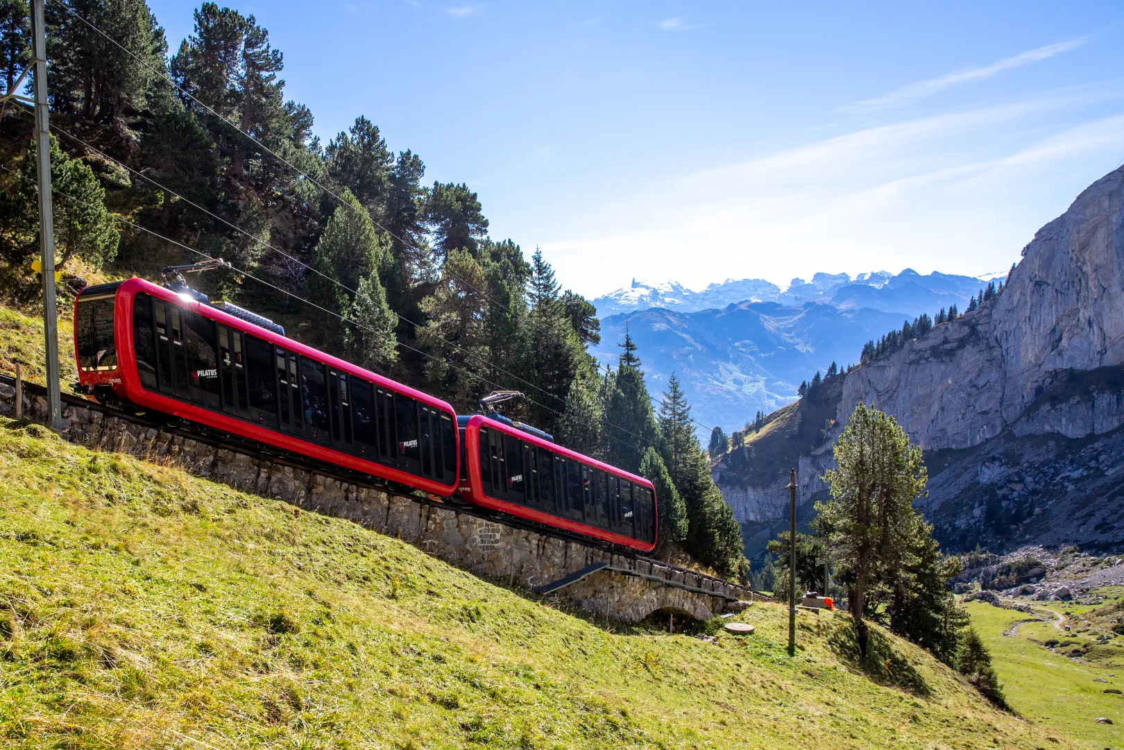 The red Pilatus cogwheel railway runs through a green landscape, with cliffs and mountain panorama in the background.