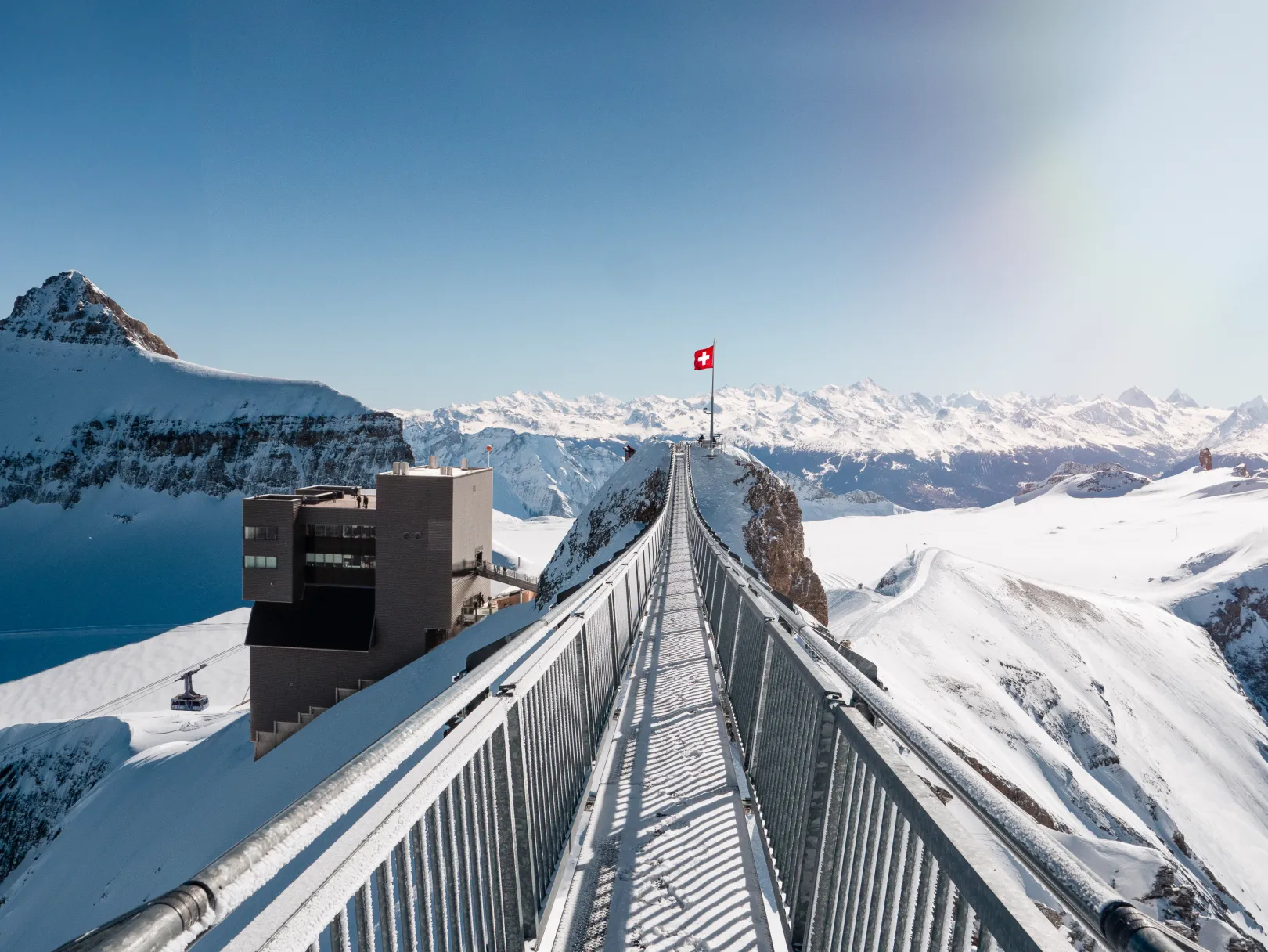 The Peak Walk by Tissot on Glacier 3000 leads to the viewpoint with the Swiss flag, surrounded by snow-covered Alps and a blue sky.