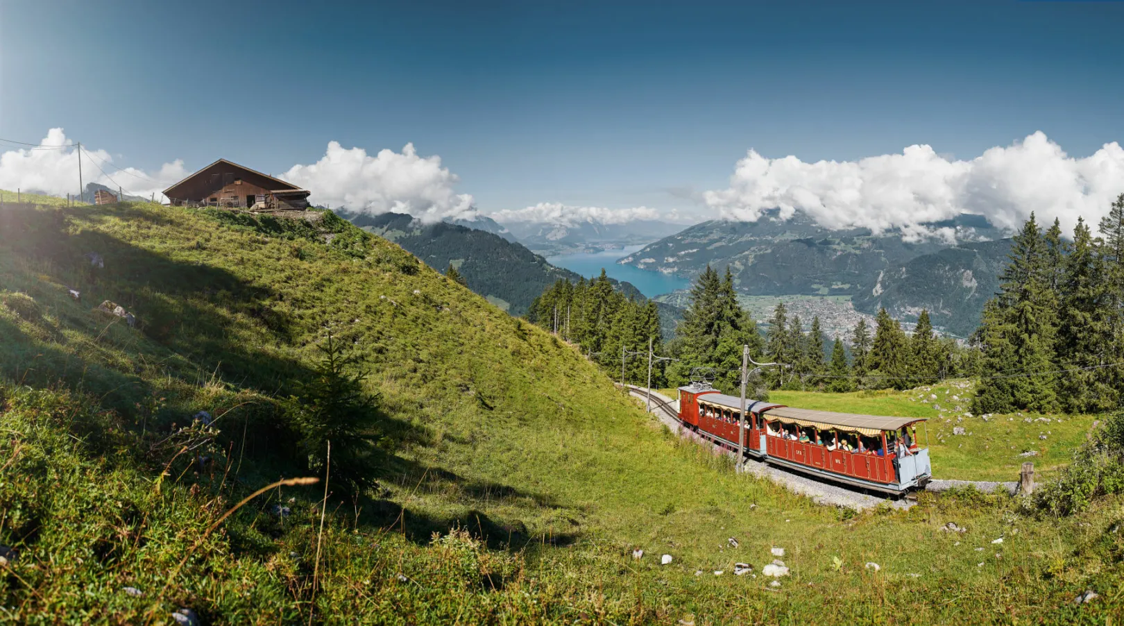 The Schynige Platte Railway runs through a green landscape, with a blue lake and forested mountains in the background.