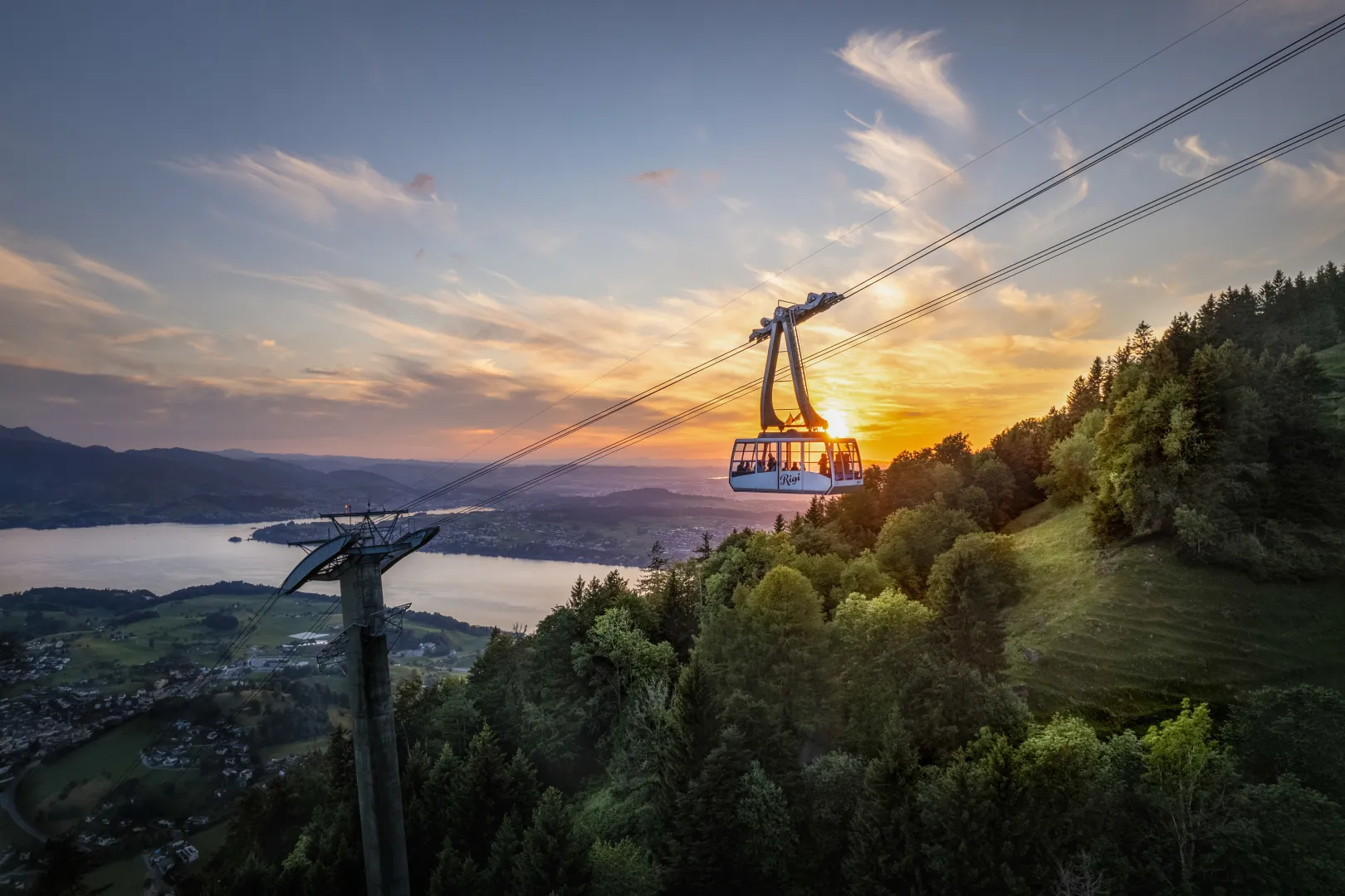 The Rigi cable car floats at sunset over wooded hills; below lies Lake Lucerne in a wide pre-Alpine landscape.