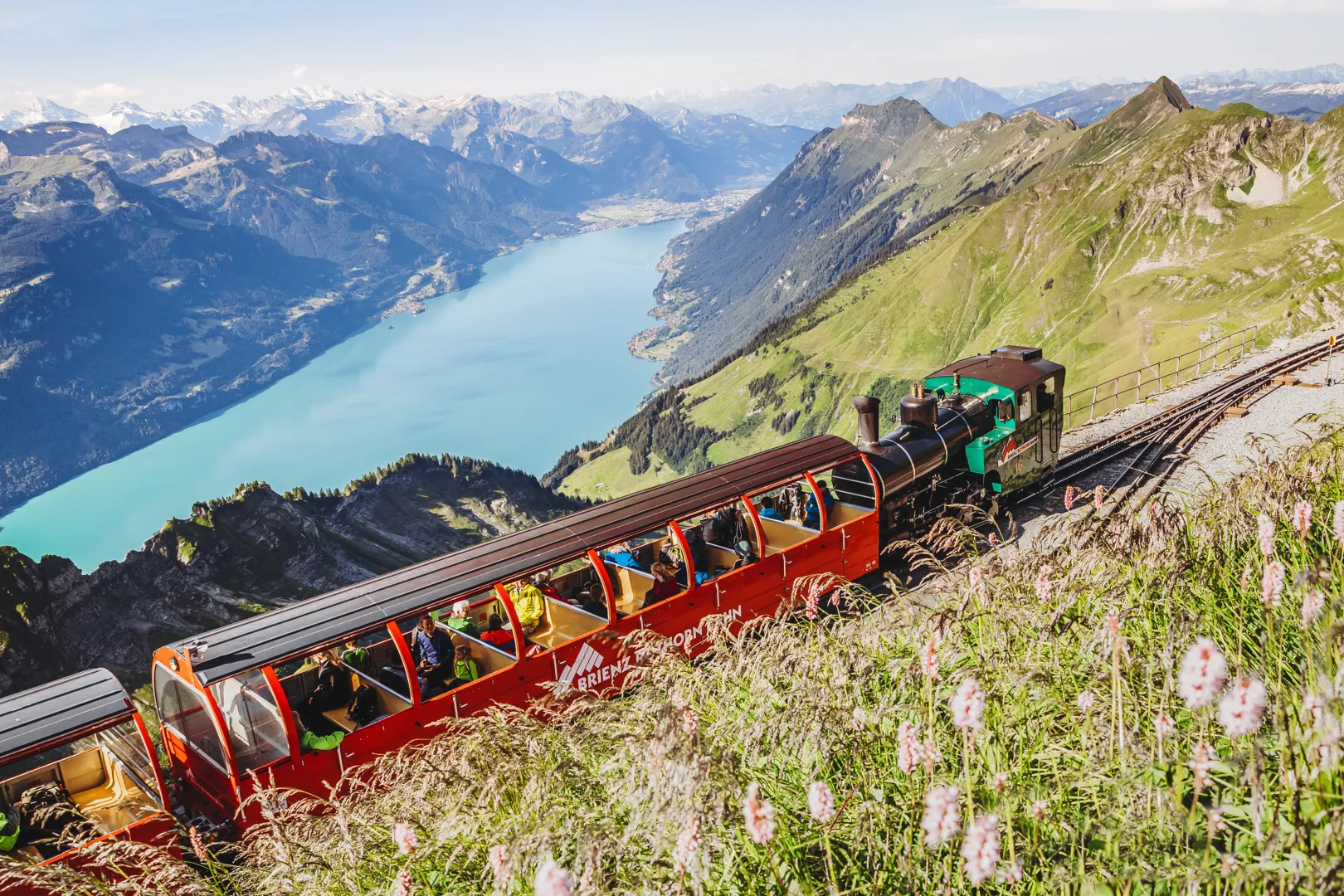 The Brienzer Rothorn Railway climbs the mountainside with open red carriages, with the turquoise Brienz Lake under a clear sky in the background.