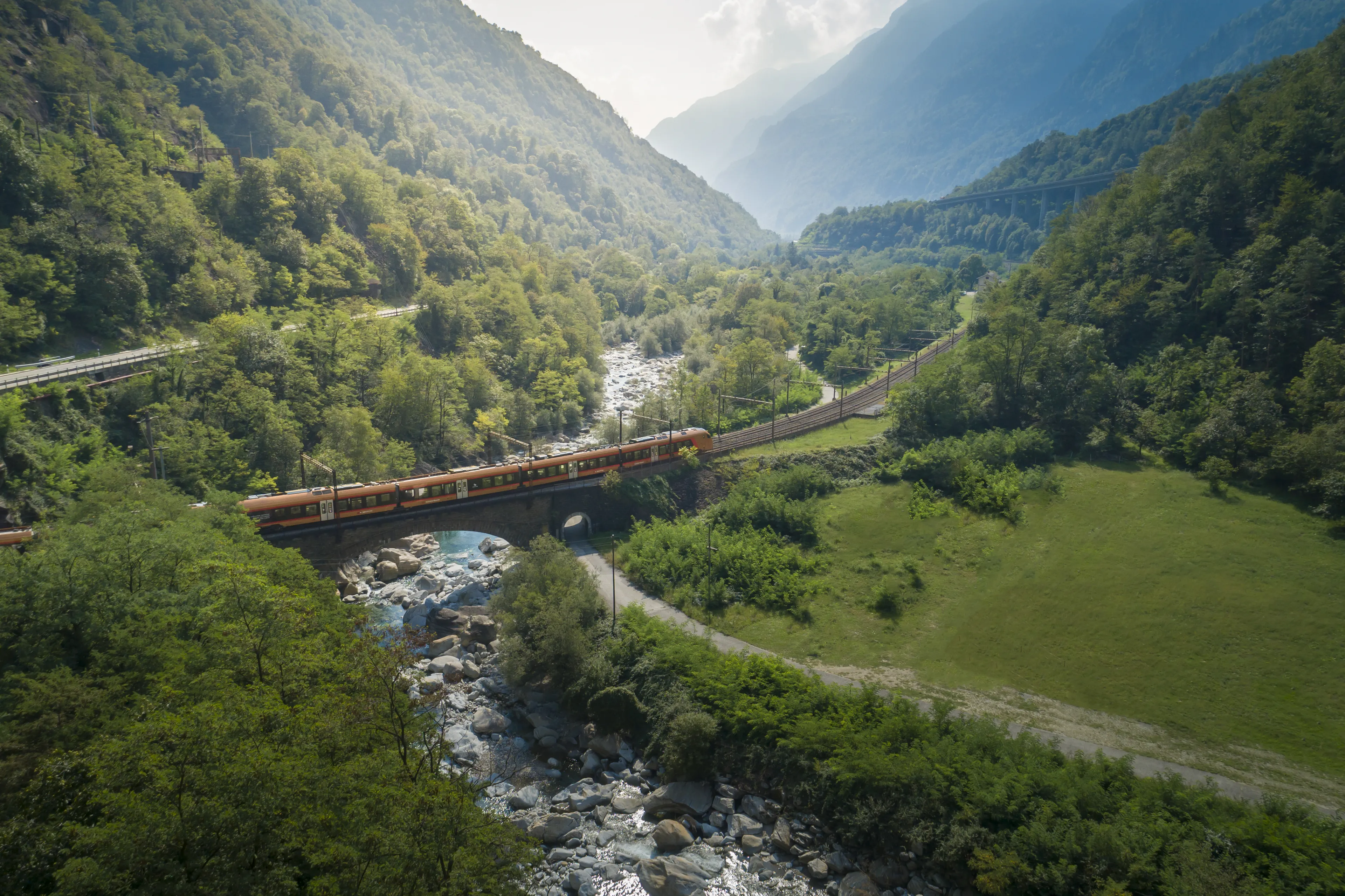The Treno Gottardo passes through green countryside near Wassen and crosses a bridge over a river.