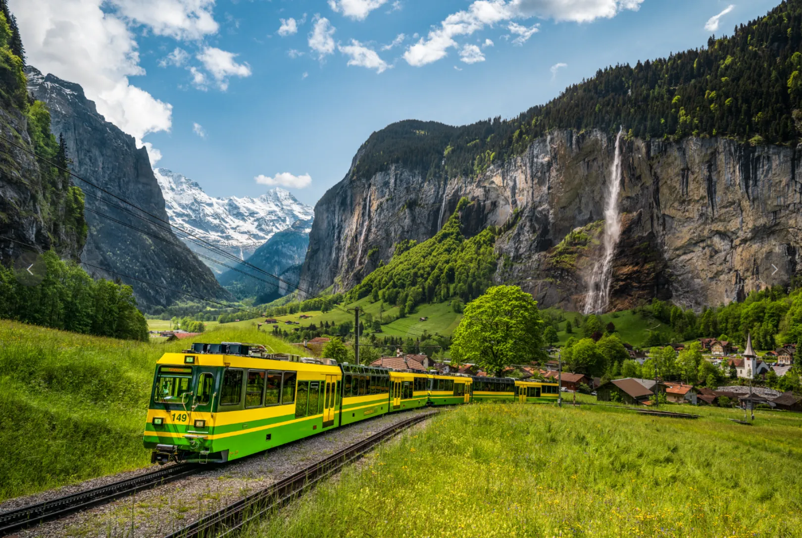 The Wengernalpbahn passes through a green meadow landscape in Lauterbrunnen, with steep rock faces, a high waterfall, and snow-covered mountains in the background.