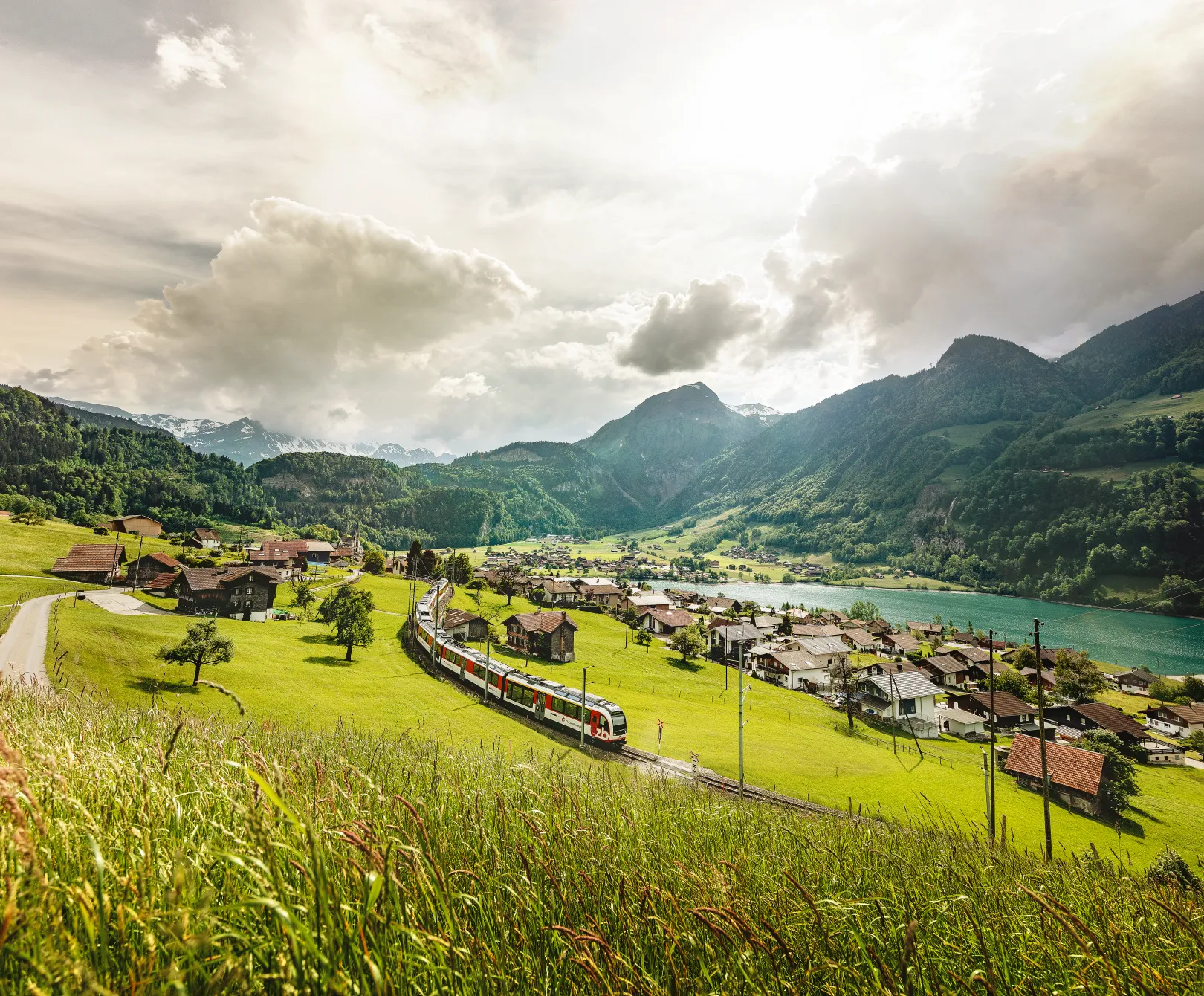The Luzern–Interlaken Express passes through a green mountain landscape in Lungern in summer, with views of a village and a lake.